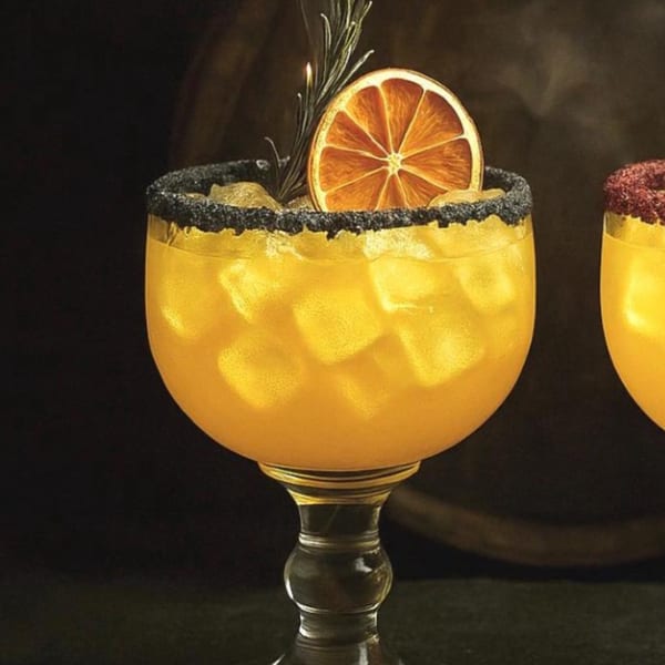 A vibrant orange cocktail with ice, a dried orange slice, and rosemary, rimmed with black salt.