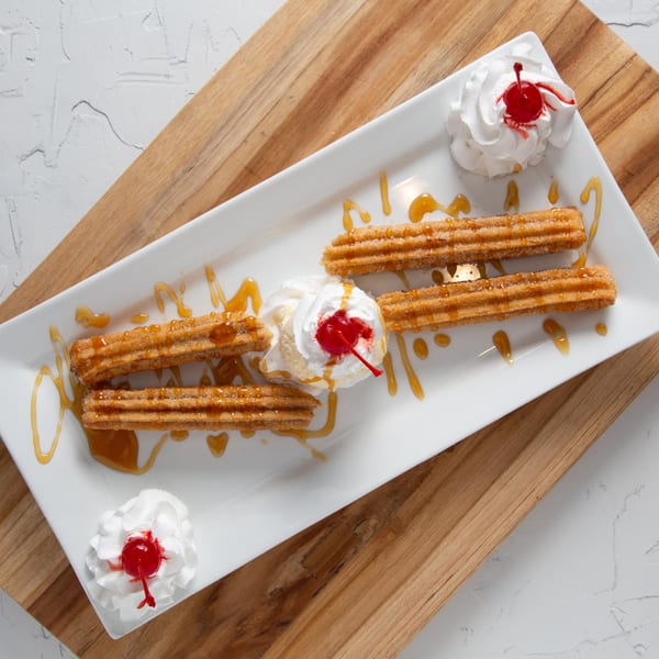 Los Compas churros with whipped cream, cherries, and caramel drizzle on a white plate.